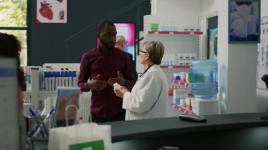 Afro-Amerikalı bir adam eczaneden ilaç ve vitamin almak için ilaç tedavisi hakkında uzman birini arıyor. Genç bir yetişkin, yaşlı bir kadınla sağlık sigortası hakkında konuşuyor..