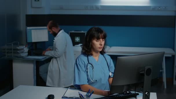 检查期间 身穿蓝色制服的医疗助理深夜在诊所办公室的电脑上输入药物治疗 下班后在医院工作的疲惫护士 医学概念 — 图库视频影像