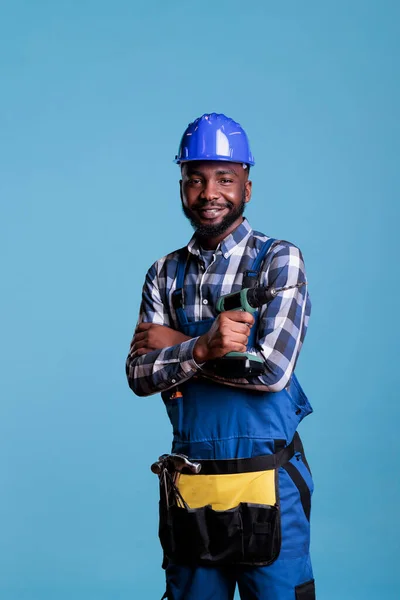 Professional contractor with hard hat and tool belt on light blue background. African american builder holding cordless drill wearing utility waist band while looking smiling at the camera.
