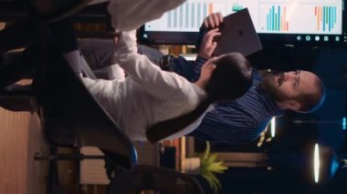 Dikey video: Şirketin gelir raporunu sunan adam, dizüstü bilgisayar ekranı ve dijital panodaki verileri karşılaştırıyor. Araştırma istatistikleri görselleştirme, analiz sunumunu dinleyen çeşitli takım