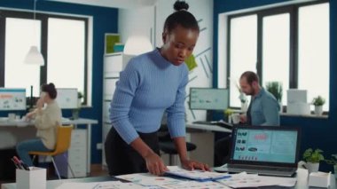 Busy african american in startup putting clipboard on desk looking at papers with sales charts comparing results with data on laptop screen. Business analyst searching for statistics while typing.