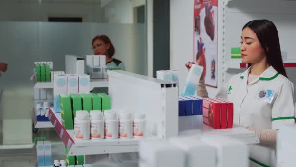 ドラッグストアで薬剤師として働いている若い女性は 棚の上の薬のパッケージを見て サプリメントの薬局のアシスタントチェックボックスと医療のお客様を助ける — ストック動画