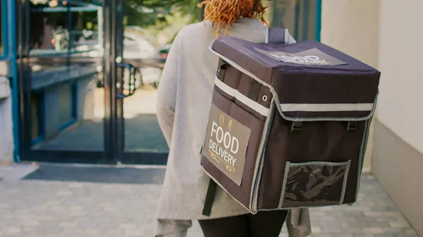 ヘッドフォンでアフリカ系アメリカ人の宅配便は 徒歩で食べ物を届け 音楽を聴き 正面玄関を歩く 持ち帰り用のバックパックから食事箱のパッケージを提供する準備 — ストック写真
