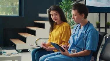 Hastane lobisinde Asyalı kadına danışmanlık yapan bir tıp asistanı dijital tablet hakkında notlar alıyor ve raporları dolduruyor. Hemşire bekleme odasındaki hastaya sağlık kontrolü yapıyor..