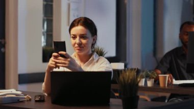 Akıllı telefondan mesaj atan mutlu ofis çalışanının portresi, dizüstü bilgisayardaki finansal paylaşım verileriyle çalışmak için yatırım uygulamasını kullanıyor. Başlangıç gelir raporu gece geç saatlerde planlanıyor.