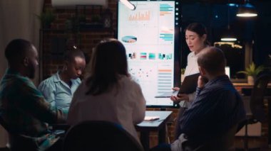 Asyalı çalışan, şirketin gelir raporunu sunuyor, dizüstü bilgisayardaki grafikleri gösteriyor. Dijital panoda istatistiksel görselleştirme, analiz sunumunu dinleyen çeşitli takımlar. Üçayak atışı.