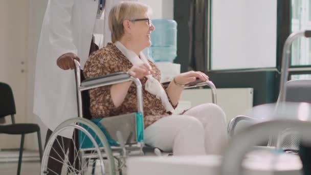 保健所で歩行フレームを使用して車椅子のシニア女性 治療を受けた後 健康的な感じ 男性医師がロビーにいる高齢者を少女と会うために連れてくる 手持ち撮影 — ストック動画