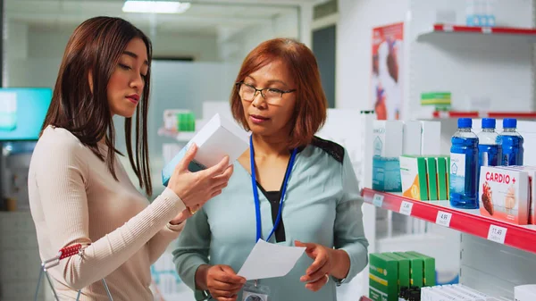 アジアの女性に治療や薬を与えるために薬の専門家の読書処方紙 医薬品店からサプリメント 薬やビタミンボックスを購入しようとしているクライアント — ストック写真