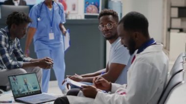 Bir grup erkek, bekleme alanındaki bilgisayardaki web kamerasını kullanarak doktorla görüntülü görüşme yapıyor. Doktor ve hasta resepsiyonda oturup teletıp çağrısında doktorla konuşuyorlar..