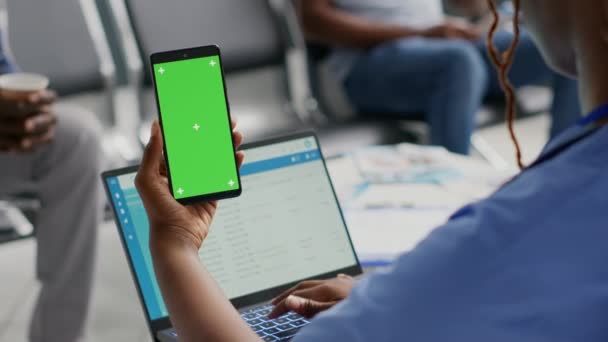 病院の受付でクロマキーディスプレイを使用して 緑の画面でスマートフォンを保持アフリカ系アメリカ人女性 孤立したコピースペースを見ている人モックアップの背景 待合室 — ストック動画