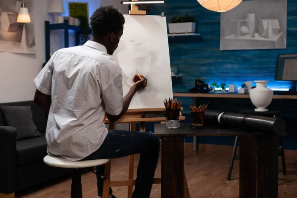 芸術的能力を持つアフリカ系アメリカ人は 花瓶の鉛筆画に仕上げを入れています 才能ある若い男作成鍋アウトライン上のキャンバスイーゼルでホームクラフトアトリエ — ストック写真
