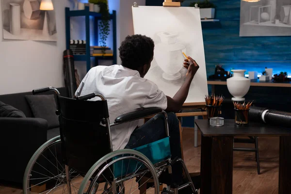 Young african american male artist user of wheelchair having good time making pencil drawing on canvas easel of pot. Man with artistic skills creating artwork at home art studio.