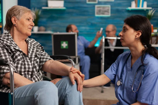 stock image Female nurse supporting elderly woman with disability in nursing home waiting room. Male nurse taking temperature of senior patient with infrared thermometer, seated on couch in background.