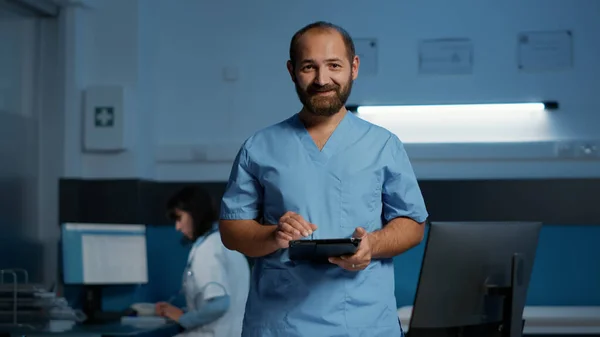 stock image Physician nurse holding tablet computer checking patient diagnosis while typing medical expertise during night shift in hospital office. Assistant man in blue uniform working at health care treatment