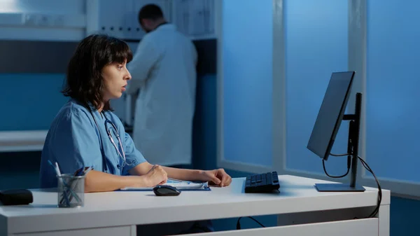 医务助理在医院办公室上夜班时 在电脑上检查病人的病情报告 以帮助治疗疾病 执业护士研究具专业知识的论文 — 图库照片