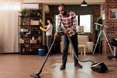 Kadın ev arkadaşı arka planda rafların tozunu alırken Afrikalı Amerikalı adam evdeki yerleri süpürüyor. Şehir dairesini temizlerken lastik eldiven takan kadın, temizlik günü..