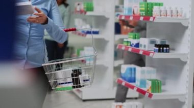 Raflarda reçeteli ilaç kutusu arayan, takviye malzemeler tutan ve broşür okuyan biri. Eczanedeki ilaç ürünlerine, sağlık ilaçlarına bakan Asyalı müşteri.