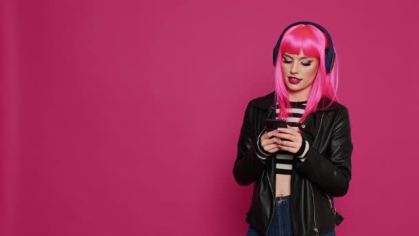 ピンクのウィッグのテキストメッセージを持つ幸せな美しい女の子は ヘッドセット上の音楽を聞いて スタジオでスマートフォン上のソーシャルメディアのオンラインアプリを使用して Mp3曲で楽しんでクールなスタイリッシュな女性 — ストック動画