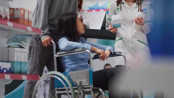 坐轮椅的妇女接受药剂师的帮助 希望购买维生素和补充剂 残疾顾客与男性护理员一起来到药店 需要药物 手持射击 — 图库视频影像