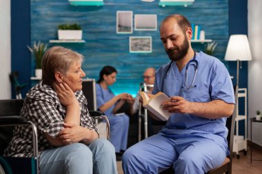Huzurevinde sosyal hizmet görevlisi tekerlekli sandalyedeki bir kadın için kitap okuyor. Yaşlı kadın huzurevi sakini erkek tıbbi asistanı dikkatle dinliyor..