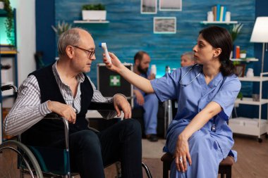 Uzman bir sağlık merkezinde hemşire tekerlekli sandalyedeki yaşlı bir hastanın ısısını dijital kızılötesi termometre kullanarak ölçüyor. Emekli adam uzmanlarla tıbbi görüşmelere katılıyor..