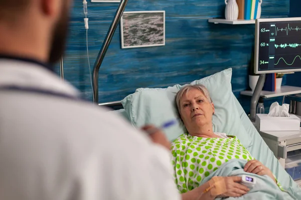 病院のベッドに寝そべっている高齢女性には男性医師が出席する 入院中の女性患者への健康診断と治療の結果を専門家が説明する — ストック写真