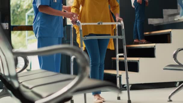 看護師は アジアの女性が歩くフレームで歩くのを助け 身体障害や障害に苦しんでいます 骨折や外傷の後に回復足の痛みを持つ患者を負傷 手持ち撮影 — ストック動画