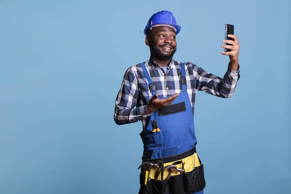 非洲裔美国人承包商在录音室拍摄的蓝色背景的手机视频通话 穿着工作服的建筑工人兴高采烈地通过智能手机谈论新的工作合同 — 图库照片