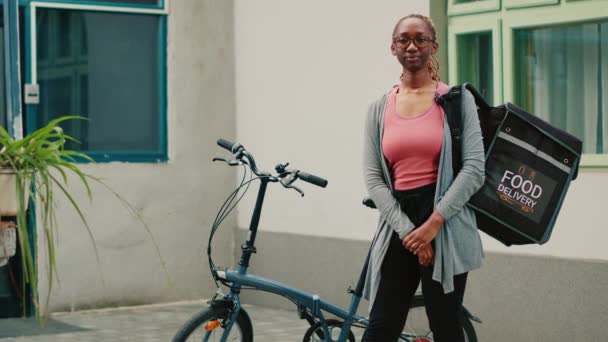 若い女性は自転車でキャリアとして働いています 紙袋に食事パッケージを提供するためにバックパックを使用して 女性は玄関でファーストフードの注文を与えるために宅配便に乗る自転車に乗る 手持ち撮影 — ストック動画