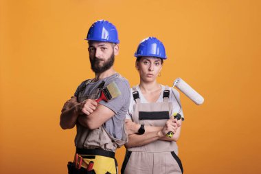 Boya aletleriyle poz veren profesyonel tamirciler, sarı arka planda duruyorlar. Boya fırçası ve tekerlekli fırça tutan inşaat ekibi stüdyoda yenileme projesi üzerinde çalışacak..