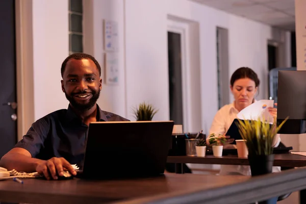 同僚のスペースの肖像画でラップトップで働く笑顔の従業員 夜にオフィスの職場の机でカメラを見ている企業の労働者 アフリカ系アメリカ人の男性がコンピュータソフトウェアを使って — ストック写真