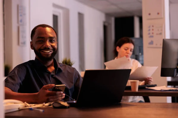 ノートパソコンで働く従業員の笑顔 コワーキングスペースのポートレートにスマートフォンアプリを使用することで プロジェクトマネージャーは職場のデスクでカメラを見ています アフリカ系アメリカ人男性がコンピュータに関するレポートを書く — ストック写真