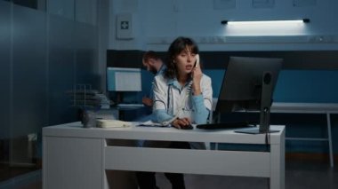 Uzaktan kumandalı bir hastayla konuşurken masasında oturan pratisyen kadın sabit telefon kullanarak ilaç tedavisini tartışıyor. Sağlık personeli gece geç saatlere kadar hastane ofisinde çalışıyor.