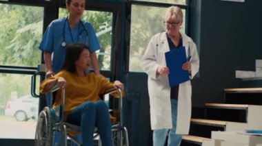 Yaşlı doktor tekerlekli sandalye kullanıcısıyla konuşuyor ve bekleme odasında oturan yaşlı Asyalı kadına danışıyor. Doktor, hastası olan hastalara destek ve tavsiye veriyor. El kamerasıyla..