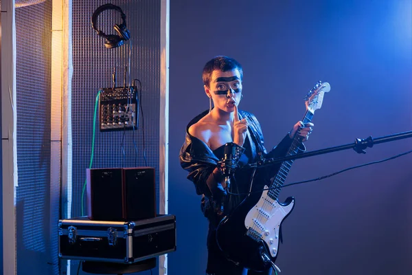 Musical performer doing hush mute sign, playing guitar at microphone and showing secret privacy gesture. Heavy metal singer keeping silence and secrecy, confidential symbol in studio.