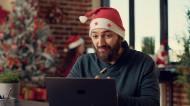 Erkek işçi web kamerasıyla video konferansında konuşuyor, Noel arifesinde Noel Baba şapkası takıyor. Xmas tatil süslemeleriyle internette sohbet eden bir adam..