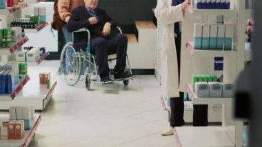 Hasta bakıcı ve yaşlı adam eczanede tedavi arıyorlar, ilaçlarla ilgili uzmanla konuşuyorlar. Tekerlekli sandalye kullanan engelli, bakıcı ilaç istiyor..