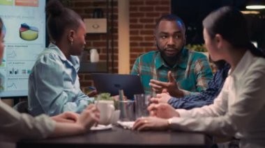 İş toplantısı, Afro-Amerikan analitik uzmanı şirketin gelir raporlarını dizüstü bilgisayardan gösteriyor. İş arkadaşları istatistik sunumunu tartışıyor, çalışanlar analistleri dinliyor.