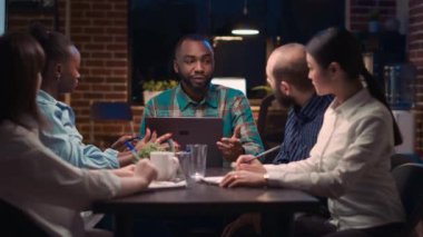 Afrika kökenli Amerikalı analitik uzmanı iş toplantısında laptop ekranında veri görselleştirmesi gösteriyor. Takım çalışması, çeşitli iş arkadaşları şirket raporlarını gece vakti tartışıyor.