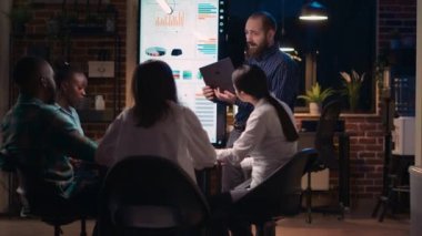 Şirketin gelir raporunu sunan adam dizüstü bilgisayar ekranı ve dijital panodaki verileri karşılaştırıyor. Araştırma istatistikleri görselleştirme, iş toplantısında analitik sunumu dinleyen çeşitli takım