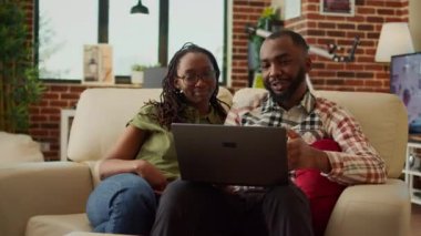 Afro-Amerikalı çift dizüstü bilgisayardan video izliyorlar, kanepede dinleniyorlar ve eğleniyorlar. İnternette gezinmek, boş zamanların tadını çıkarmak, evde gülmek..