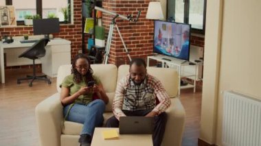 Afrikalı Amerikalı modern çift evde koltukta dinleniyor, akıllı telefondan sosyal medya uygulamalarına kayıyorlar. Eğlencenin tadını çıkarmak, birlikte gülmek, oturma odasında eğlenmek..