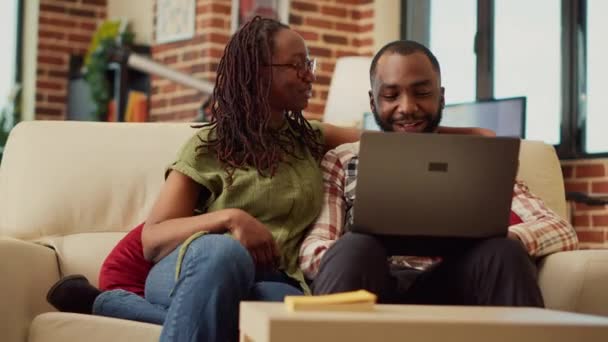 アフリカ系アメリカ人のカップルが一緒にラップトップでビデオを見て ソファに座って自宅でリラックスします レジャー活動をオンラインで楽しむ関係で若いパートナーは 笑いの家にご滞在 手持ち撮影 — ストック動画