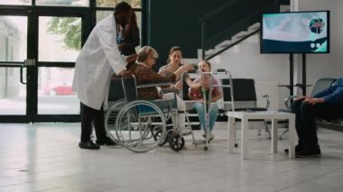 Tekerlekli sandalyedeki yaşlı bir kadın bekleme odasında küçük bir kızla konuşuyor. Ailesi yaşlı bir hastayı ziyarete geliyor. Tekerlekli sandalye kullanıcısı hastane lobisinde yürümeye çalışıyor..