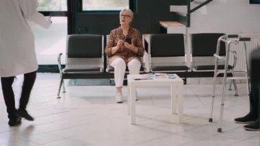 Yaşlı bir kadını muayene odasına götüren erkek sağlık uzmanı, yaşlı bir hasta bekleme salonunda oturuyor. Pratisyen hekimle check-up randevusu olan kişi.
