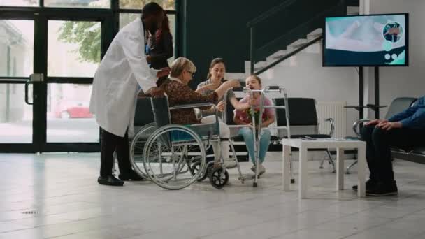車椅子の高齢女性は待合室で女の子と話をし 家族は障害のある高齢者を訪問するようになる 車椅子をご利用の方は 病院のロビーを歩いてください — ストック動画