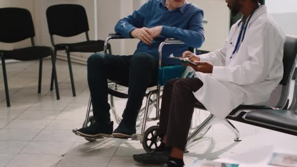 Γενικός Ιατρός Που Συμβουλεύεται Ηλικιωμένο Άτομο Αναπηρική Καρέκλα Συμπληρώνοντας Ιατρικά — Αρχείο Βίντεο