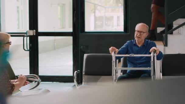 歩行フレームを持つ高齢者の患者が待機エリアに座って 医療サービスや医療について女性と話をする 病院のロビーで健康診断を受ける老人 — ストック動画