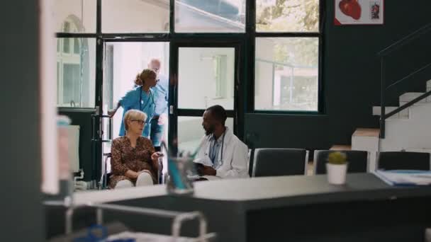 アフリカ系アメリカ人医師が車椅子で老婦人に相談し 病院の待合室で身体障害や病気について話しています 専門医及び身体障害者健康診断を行う患者 — ストック動画