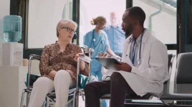 Hastahane bekleme odasında uzman biriyle konuşan yaşlı bir kadın, lobideki tekerlekli sandalye kullanıcısına danışıyor. Erkek doktor sağlık raporu alıyor..